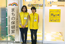 まちの保健室と書かれた旗の横に立つ、同じ黄色いベストを着た2人の女性の写真