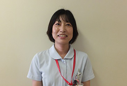 赤いストラップをかけ、薄い水色の制服を着てカメラに笑顔を向ける看護助手の女性の写真