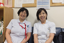 薄い水色の制服を着て並んで椅子に座る看護助手の2人の写真