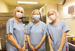 青い手術着に帽子とマスクを付けた手術室・看護助手の3人が並んでいる写真