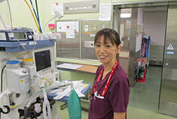 機械の前に立っているエンジ色のナースウェアを着た小林亜紀子さんの顔写真
